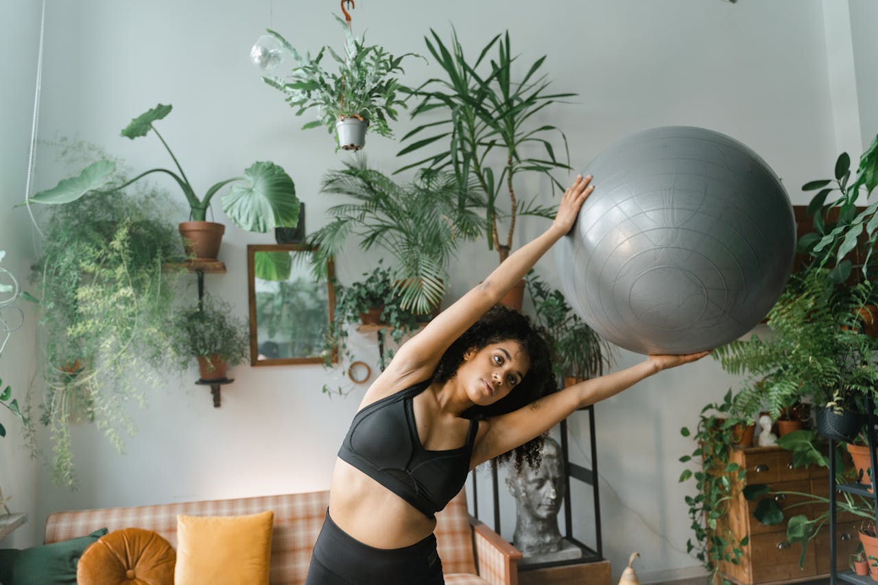 A woman stretches with a yoga ball surrounded by lush indoor plants, embracing healthy living.